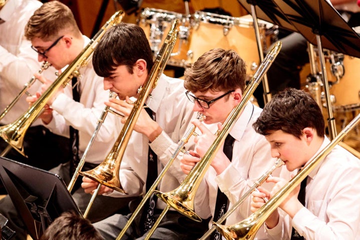Young men in white shirts in black ties are seated in a row all playing trombones.