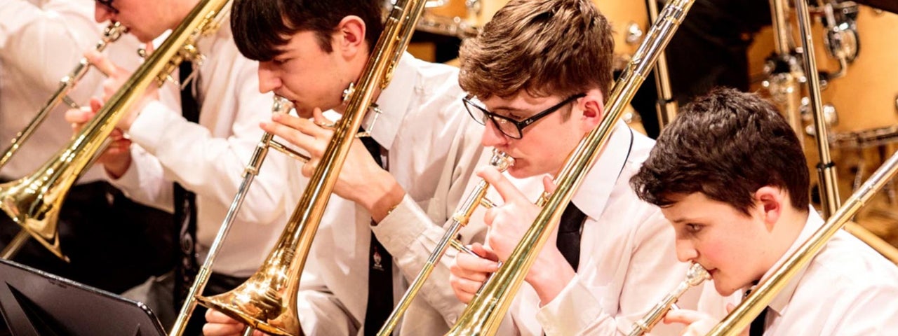 Young men in white shirts in black ties are seated in a row all playing trombones.