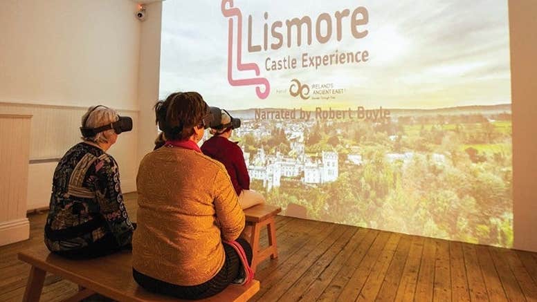 Rear view of 3 people wearing VR sets sitting on small wooden benches facing a large image projected onto a blank wall.