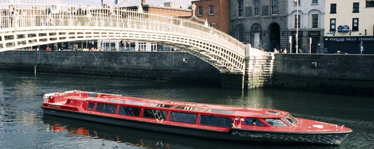 An image of the red boat cruising along the River Liffey under the Hapenny Bridge in Dublin City