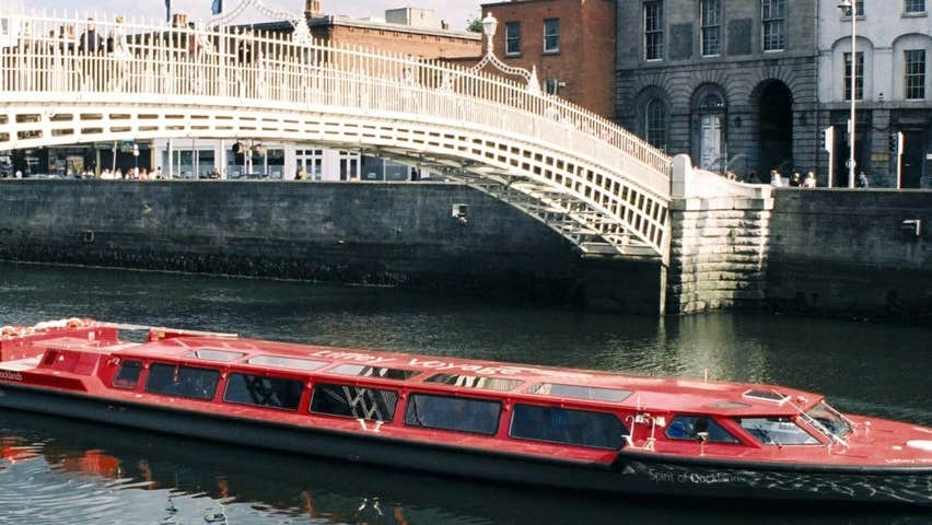 An image of the red boat cruising along the River Liffey under the Hapenny Bridge in Dublin City