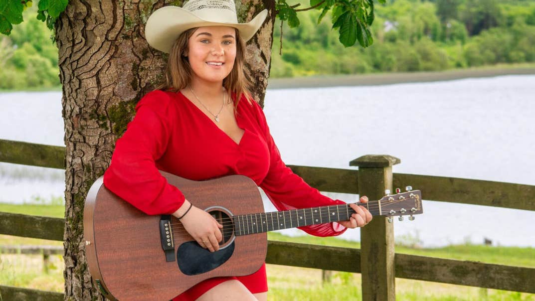 Effie Neill Dance - Gleneagle Arena, a smiling woman playing a guitar, leaning against a tree.