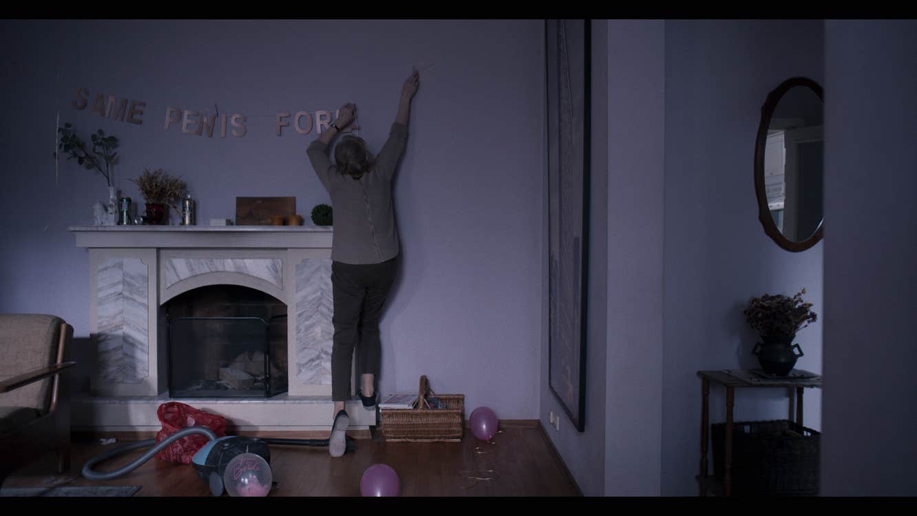 A woman hanging up letters that spell "Same Penis For" above a fireplace. The third word is apparently incomplete.