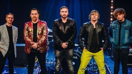 Group photo of 5 men standing in a line at the front of a stage, mostly wearing black with colourful or leather jackets, one is wearing bright lime coloured trousers, all looking at the camera, not smiling.