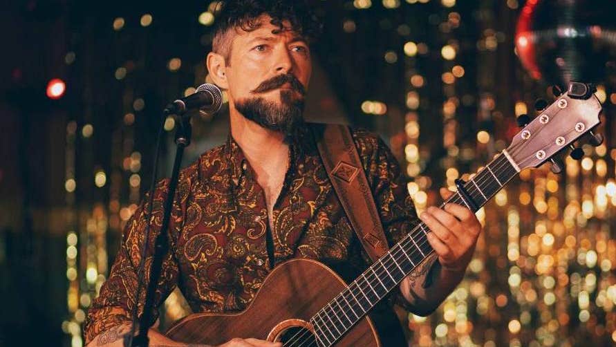 Niall McCabe to play Gleneagle Arena- a man playing a guitar standing at a mic on on stand with dark and gold blurry background.