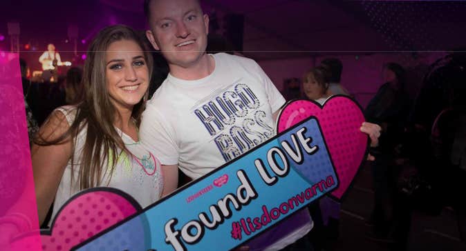2 smiling people holding pink, large hearts and a hashtag sign.