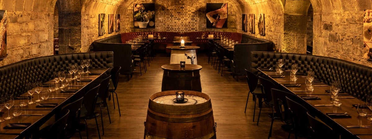 A stone walled room with a vaulted ceiling with wooden tables and beer barrels 