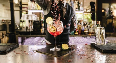 A person standing at a bar pouring liquid into a glass which is decorated with slices of fruit