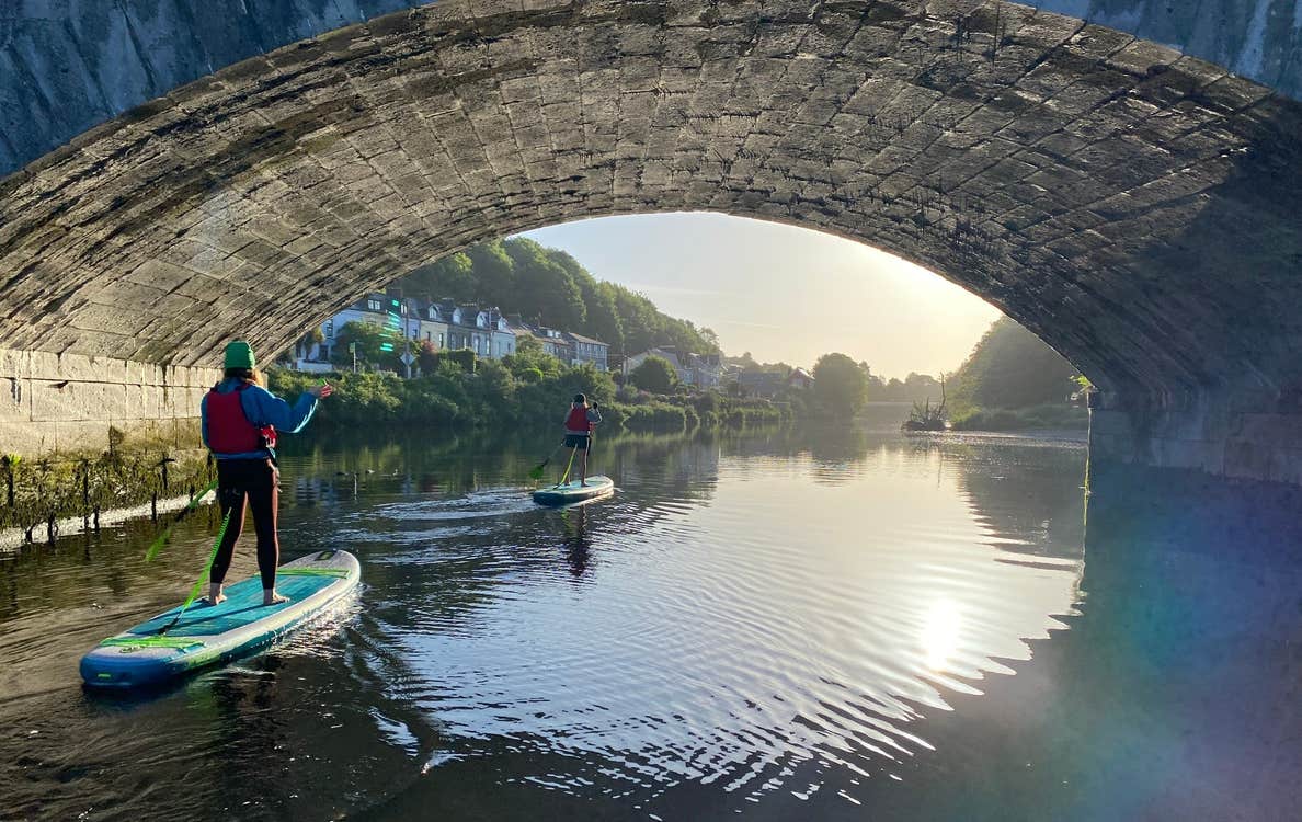 Two ladies stand up paddle boarding under a bridge with Cork City SUP