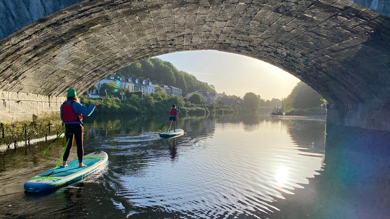 Two ladies stand up paddle boarding under a bridge with Cork City SUP