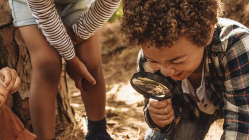 2 young children, one looking through a magnifying glasses are looking down at leafs and brown matter on the ground.