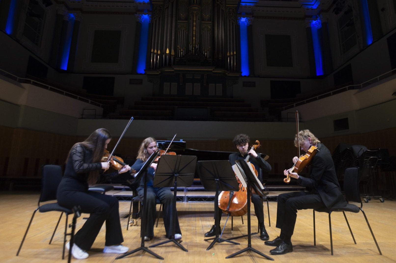 4 people dressed in dark clothing are seated in a semi circle playing stringed instruments on a large stage.