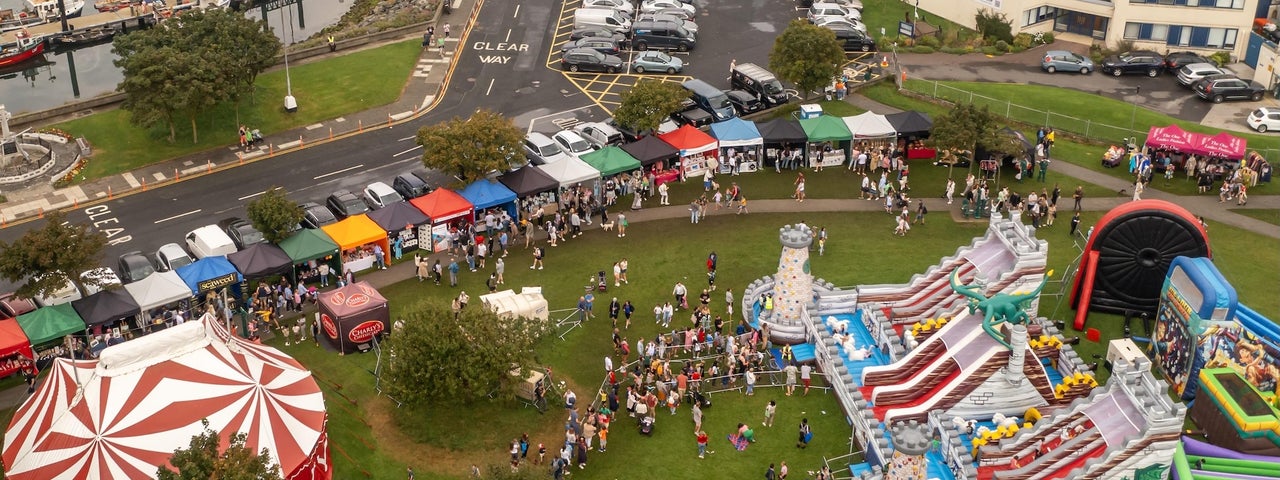 Howth Maritime & Seafood Festival 2026 - viewed from above, a grassy area containing a big top tent, large bouncy slides, rows of stalls in a town setting