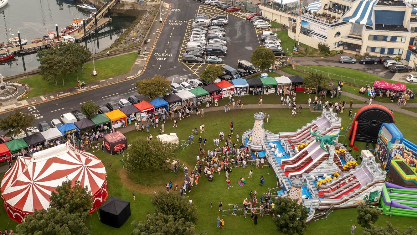 Howth Maritime & Seafood Festival 2026 - viewed from above, a grassy area containing a big top tent, large bouncy slides, rows of stalls in a town setting