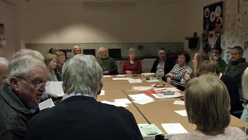 People seated around a large table with papers on listening to a woman talking.