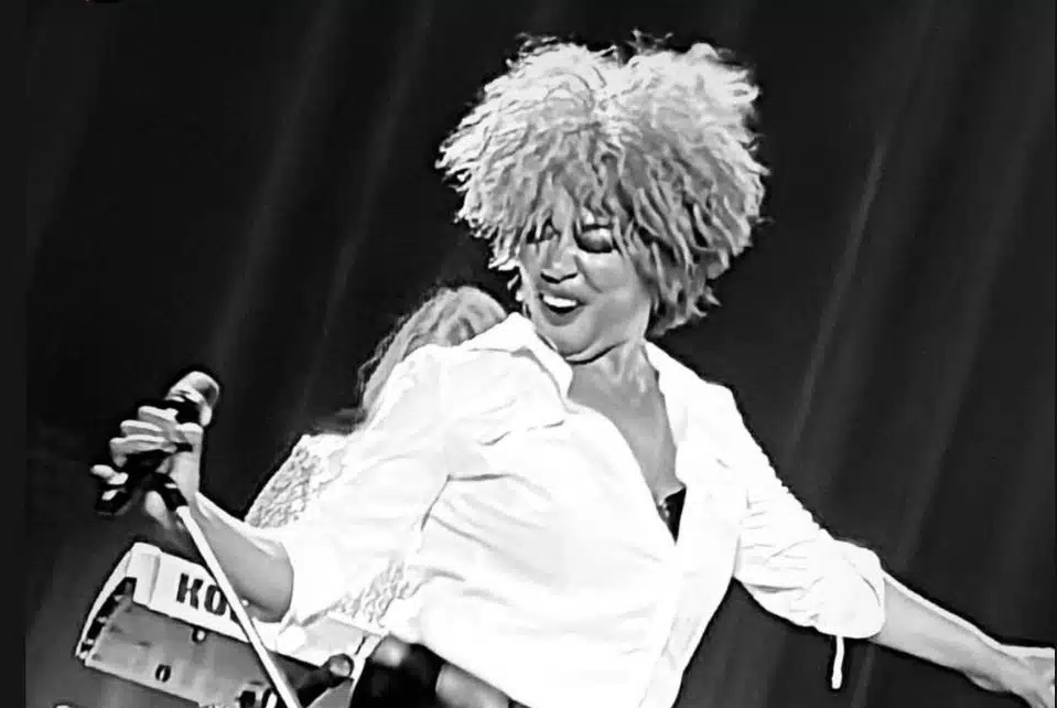 Black and white photo of woman in white shirt, performing, holding mic in right hand.