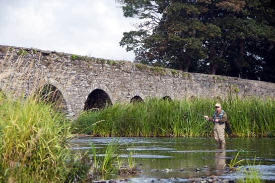 A man standing in water fishing beside an arched bridge 