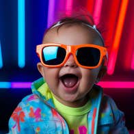 A happy baby wearing large orange sunglasses with neon tubes in background.