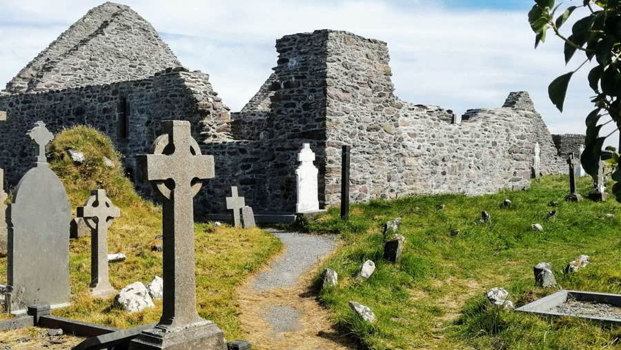 A view of the ruins of Ballinskelligs Abbey with various headstones visible in the graveyard beside it