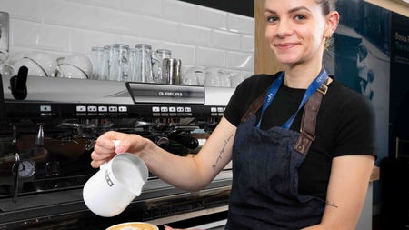 Person holding a milk frother and a cup of coffee while standing in front of coffee machine