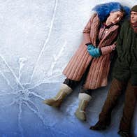 2 people dressed in winter coats are lying on their backs close together on icy surface.