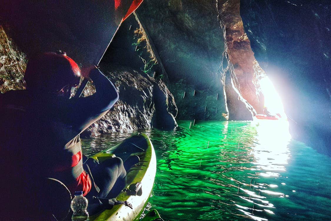 A man in a kayak in a sea cave with daylight coming through a crevice making sea water look green