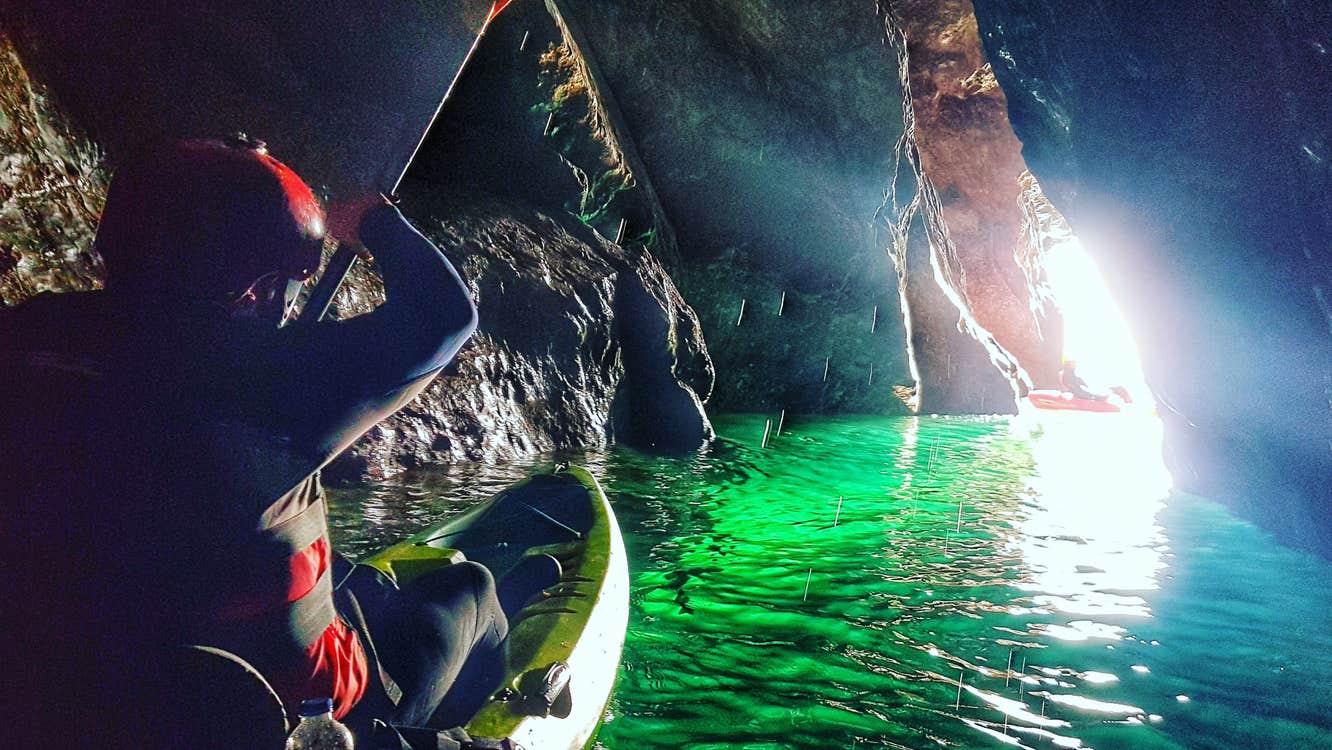 A man in a kayak in a sea cave with daylight coming through a crevice making sea water look green