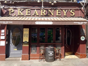 Front of pub with name over awning and two high stools beside a whiskey barrel