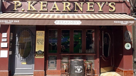 Front of pub with name over awning and two high stools beside a whiskey barrel