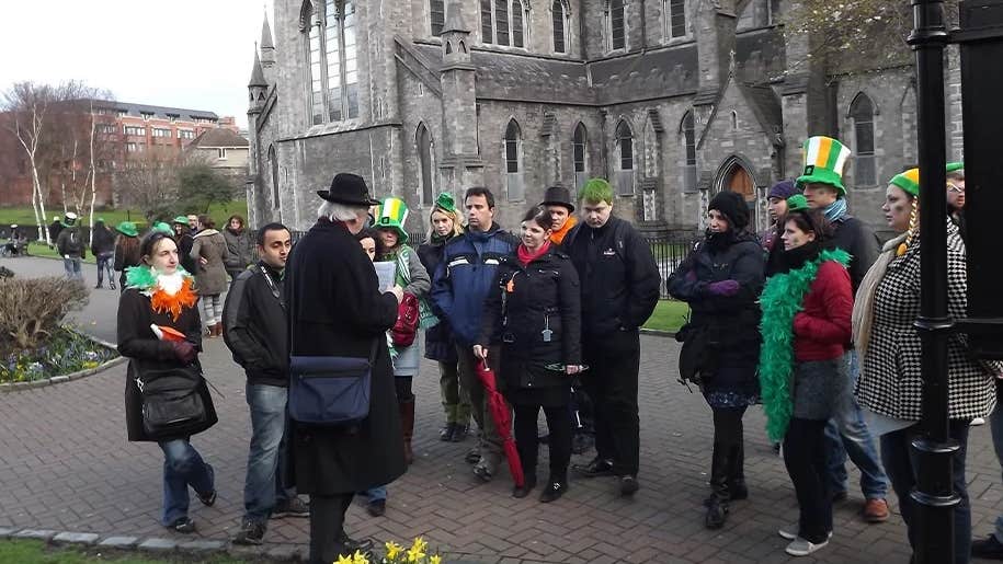 A man in dark overcoat and hat talking to a group of people outside a large, grey stone church.