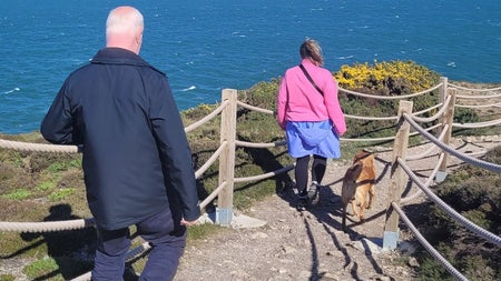 Two people walking a dog along a fenced coastal path overlooking the ocean on a sunny day