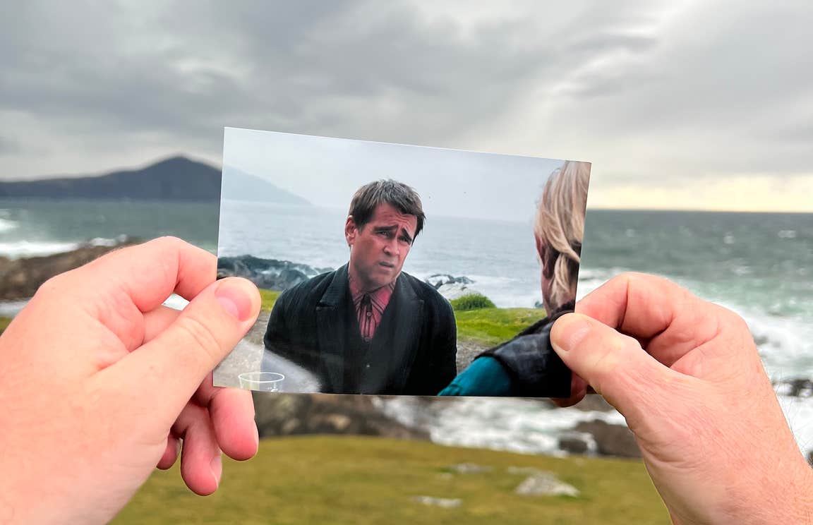 Two hands holding up a picture of a scene from the film, Banshees of Inisherin, that was filmed in that location. The scene pictures actor Colin Farrell expressing a sad face.