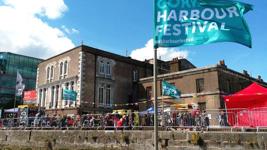 Cork Harbour Festival 2025, blue flag flying on a quay with stone buildings and people.