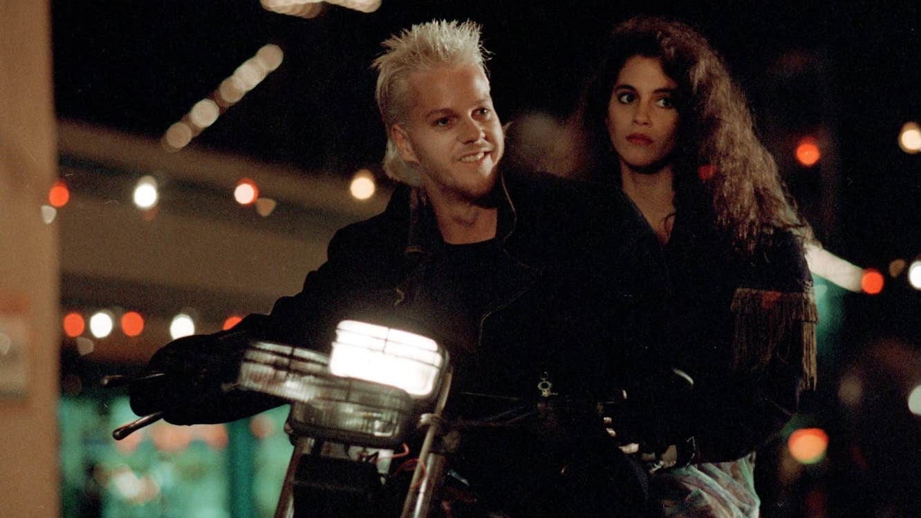 A man with a manic smile on a motorcycle. A woman with a worried expression is also on the motorcycle.