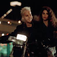 A man with a manic smile on a motorcycle. A woman with a worried expression is also on the motorcycle.