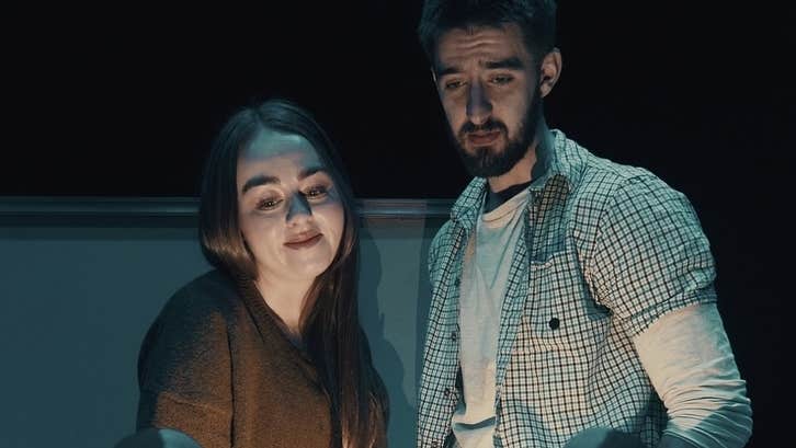 A woman and slightly anxious looking man looking down at something obscured from the viewer, dimly lit.