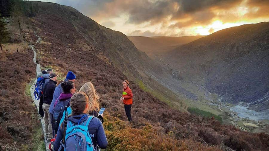 A group of hikers on a mountain trail listening to a guide as the sun sets in the distance