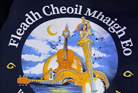 ​Fleadh Cheoil Mhaigh Eo. Circular logo including string instruments on water with blue sky.