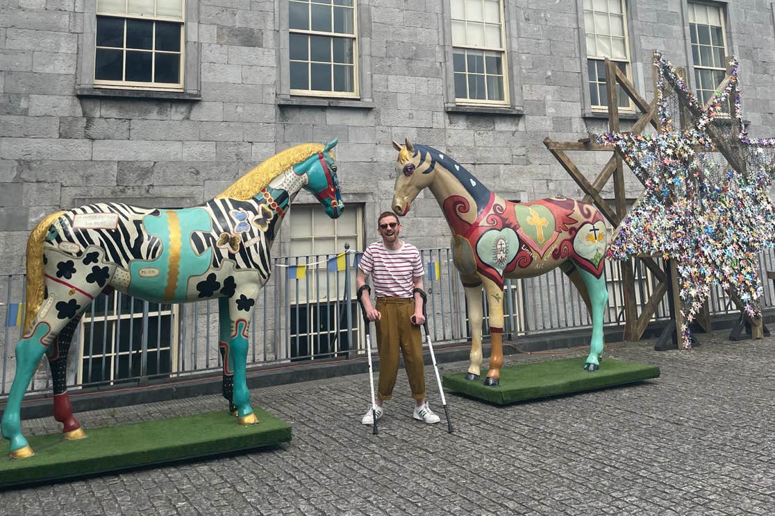 Man with crutches posing between two painted horse statues in Limerick City.