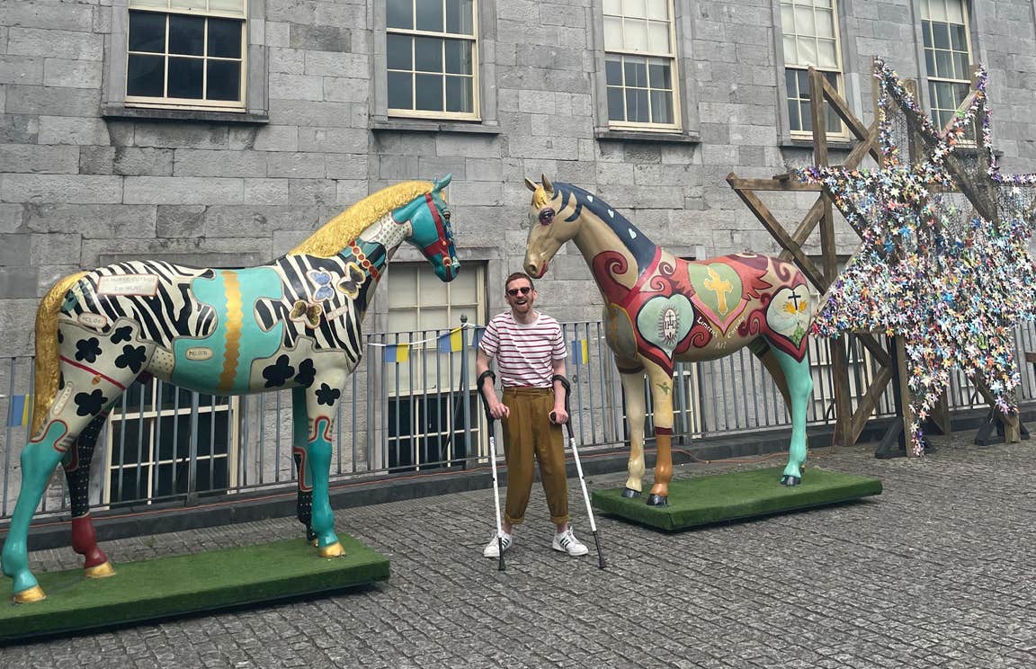 Man with crutches posing between two painted horse statues in Limerick City.