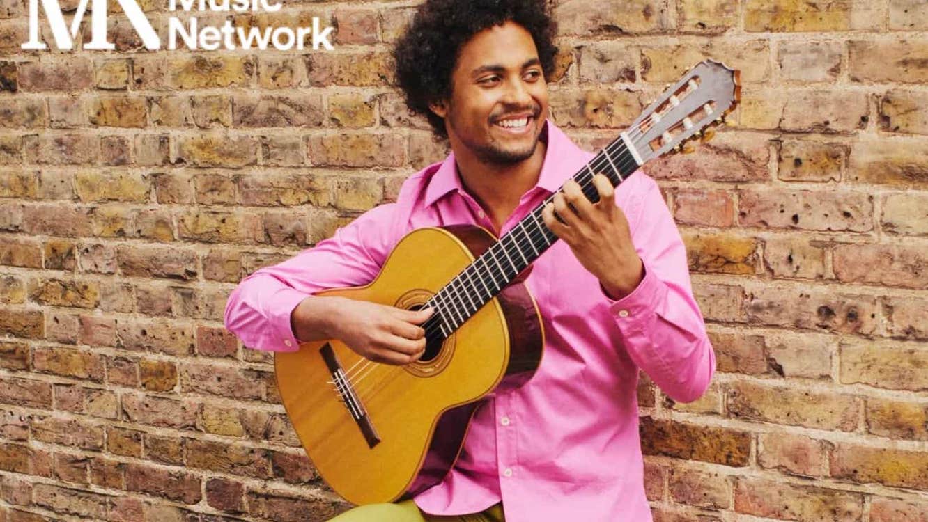A smiling man in pink shirt is playing a guitar, leaning against a yellow brick wall.