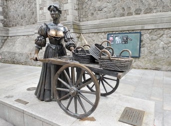 Bronze statue of a woman standing with a wheelbarrow with baskets on it