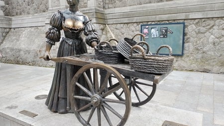 Bronze statue of a woman standing with a wheelbarrow with baskets on it