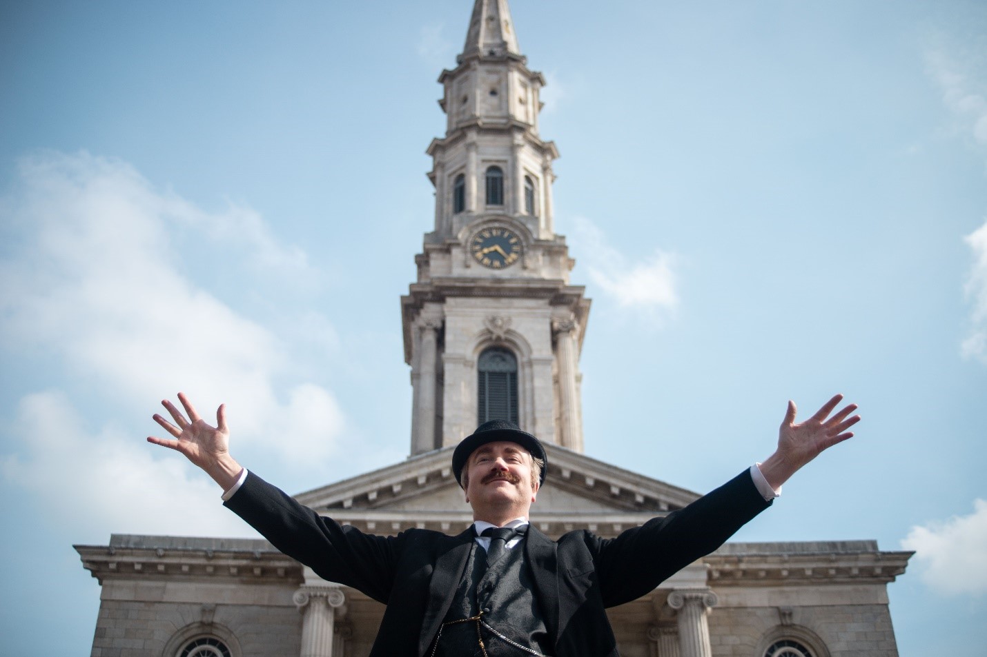 Leopold Bloom at St. George's Church Dublin. An upward shot of a man dressed in black old fashioned style clothes with his arms open wide, smiling, with the front of a church behind him and a blue sky.