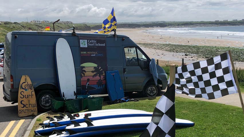 Image of Celtic Surf van parked beside the beach with surf boards and flags beside it