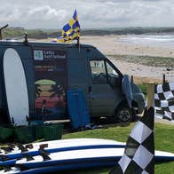 Image of Celtic Surf van parked beside the beach with surf boards and flags beside it
