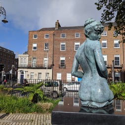 Statue of lady kneeling on black marble with a fence street cars and houses in the background