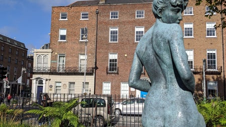 Statue of lady kneeling on black marble with a fence street cars and houses in the background