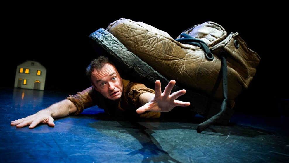 An anxious looking man is sprawled out on a stage floor reaching out with a very large boot on his back.
