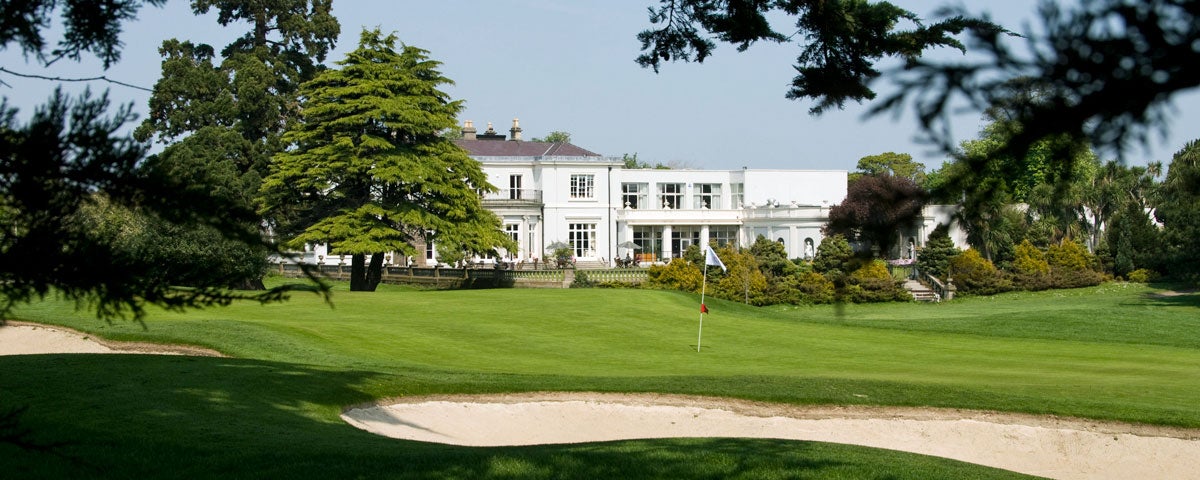 A view of Nutley House which is the name of the clubhouse in the far distance the house is two storeys high white in colour and neo Georgian in style and there is a bunker with a pinhole in the forefront of the picture